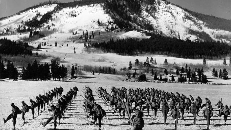Approximately 100 soldiers from the 10th Mountain Division, 87th Mountain Infantry Regiment, Company L, execute an about face on skis under the command of Lieutenant William J. Bourke on a flat snow-covered field. In the background, smaller groups of skiers are visible training in the trees at the base of one of the mountains surrounding Camp Hale, Colorado.